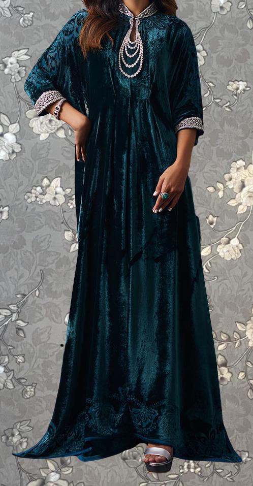 A woman wearing a long, dark green velvet dress with a high collar and sleeves, standing against a gray floral background.