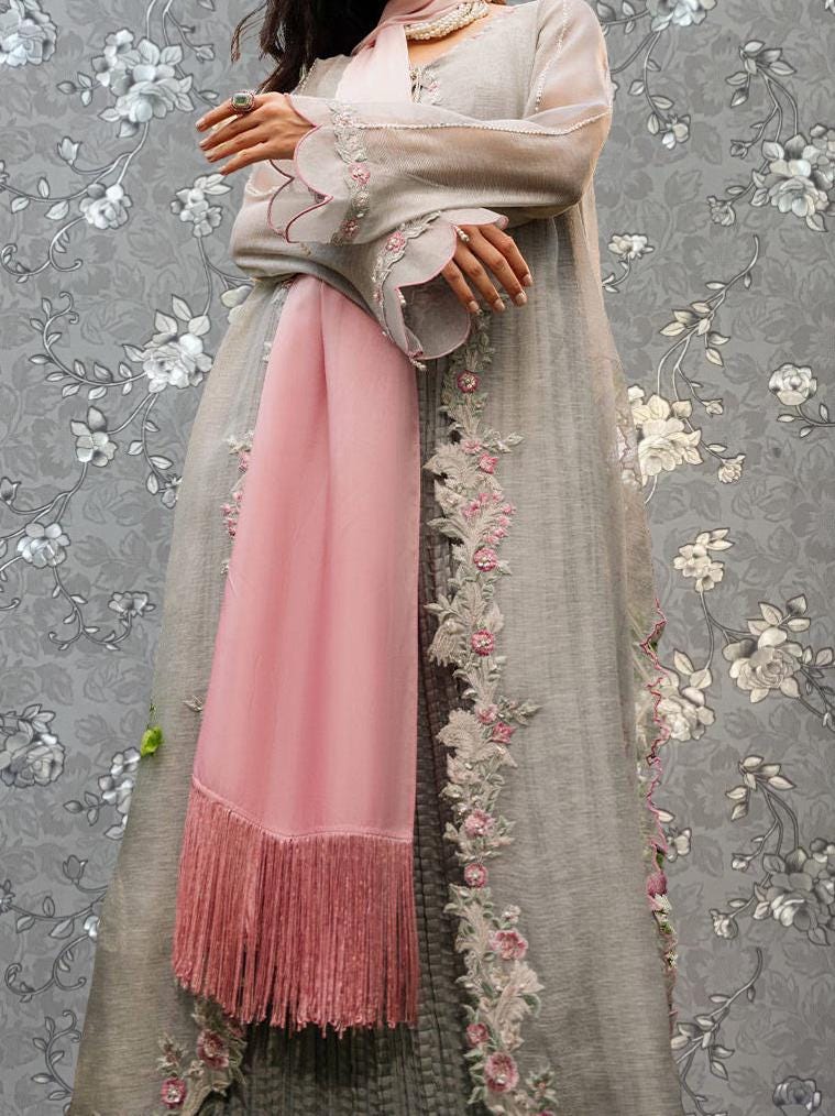 A woman wearing a long, gray dress with pink fringe and floral embroidery stands against a gray floral background.