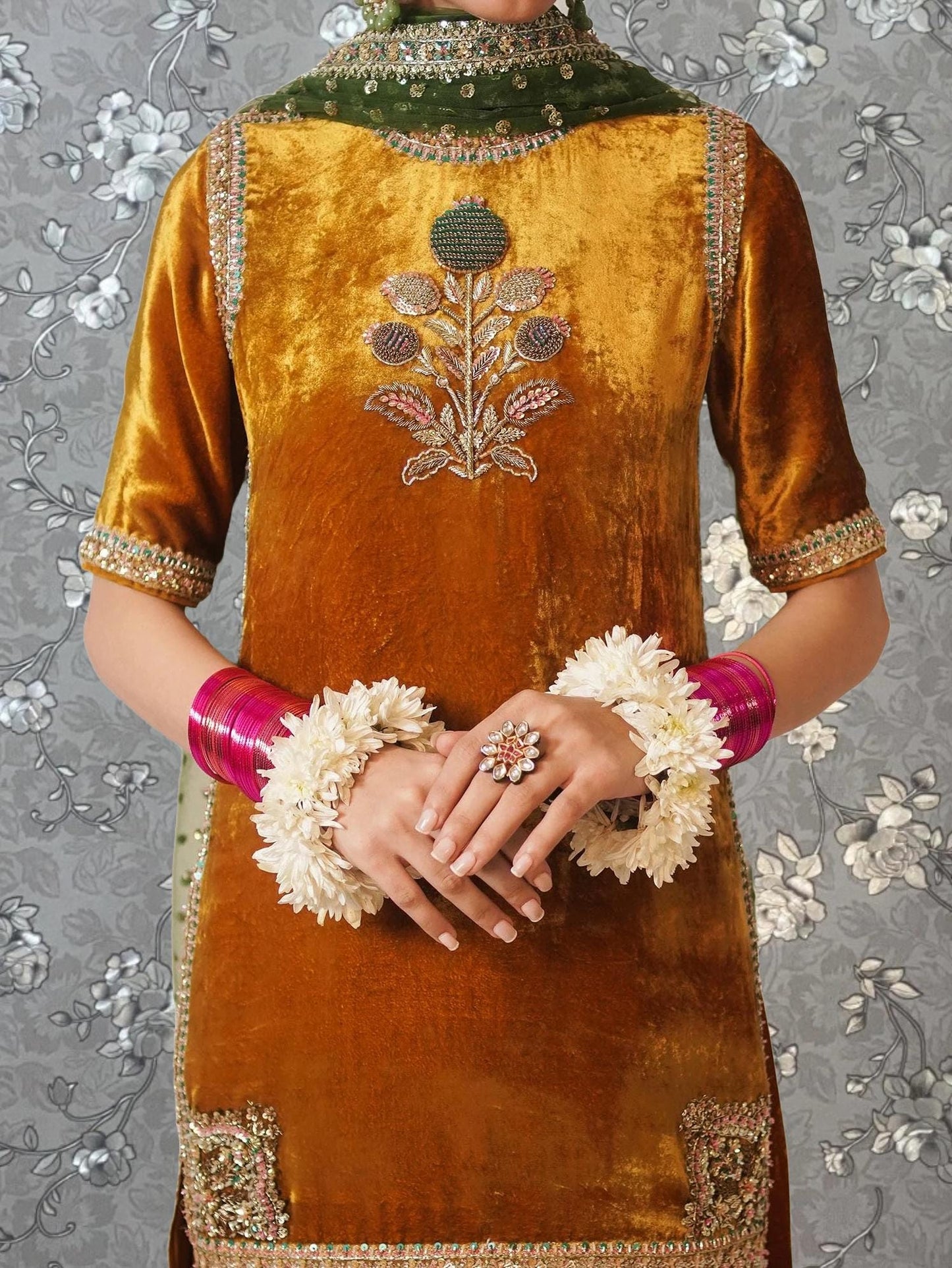 A person wearing a gold and green velvet dress with a floral design on the chest, and white flower jewelry on their hands.