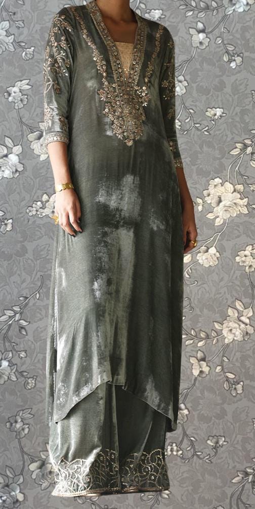 A person wearing a long, gray dress with intricate embroidery and gold accents, standing against a gray floral background.