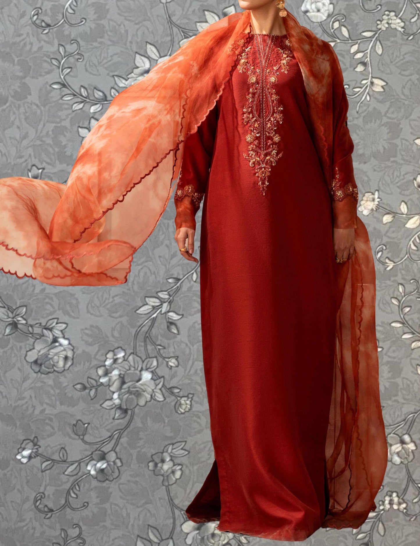A person wearing a long red dress with a flowing orange shawl, standing against a gray floral background.