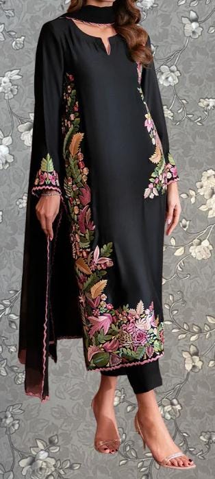 A woman wearing a long black dress with floral embroidery on the sleeves and hem, standing against a gray floral background.