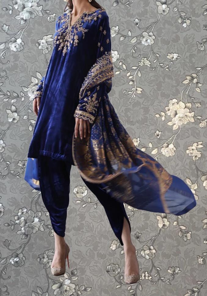 A woman wearing a blue velvet dress with gold embroidery and a matching shawl, standing against a gray floral background.