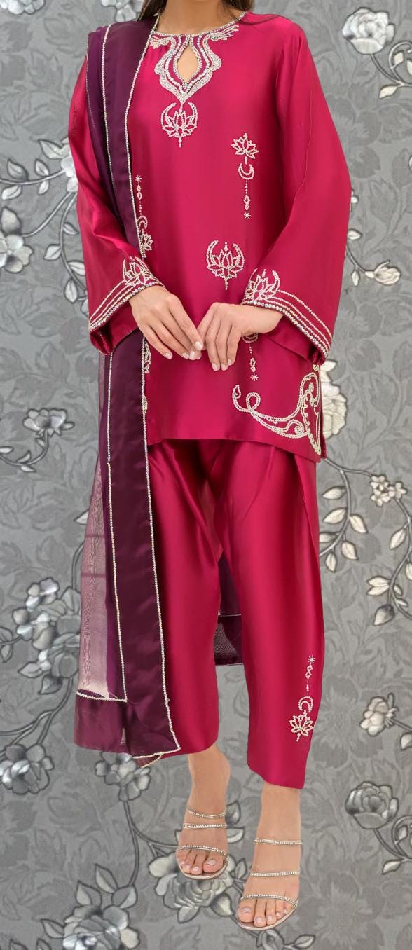 A person wearing a vibrant red outfit with intricate embroidery stands against a gray floral background.