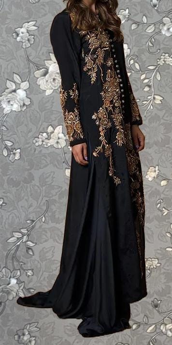 A woman wearing a long black dress with gold embroidery stands against a gray floral background.
