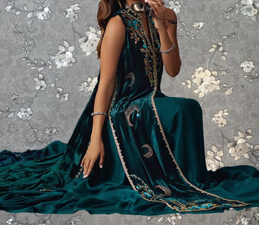 A woman wearing a long, green velvet dress with gold embroidery is sitting on a gray floral background.