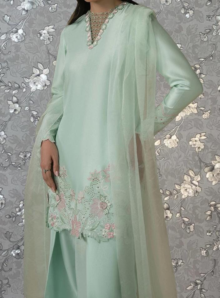 A woman wearing a mint green dress with a floral pattern and a sheer overlay, standing against a gray floral background.