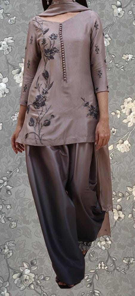 A woman wearing a brown dress with floral embroidery, standing against a gray floral background.