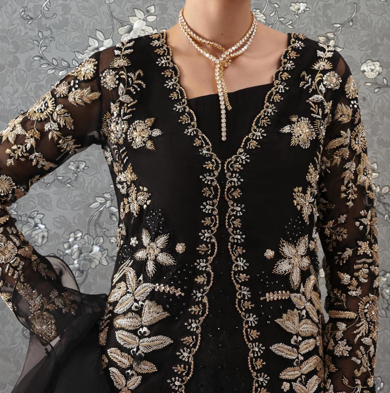 A close-up of a person wearing a black dress with intricate gold embroidery and a long necklace.