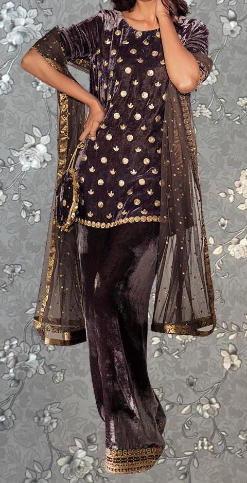 A woman wearing a purple velvet dress with gold polka dots and a matching shawl stands against a gray floral background.