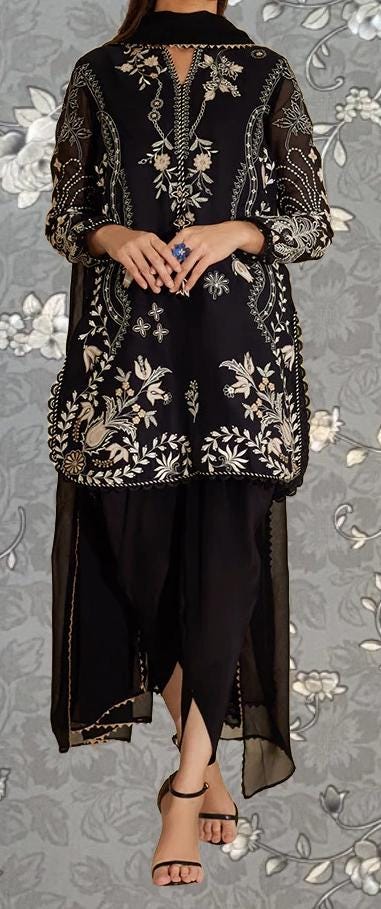 A woman wearing a black and gold dress with intricate embroidery stands against a gray floral background.
