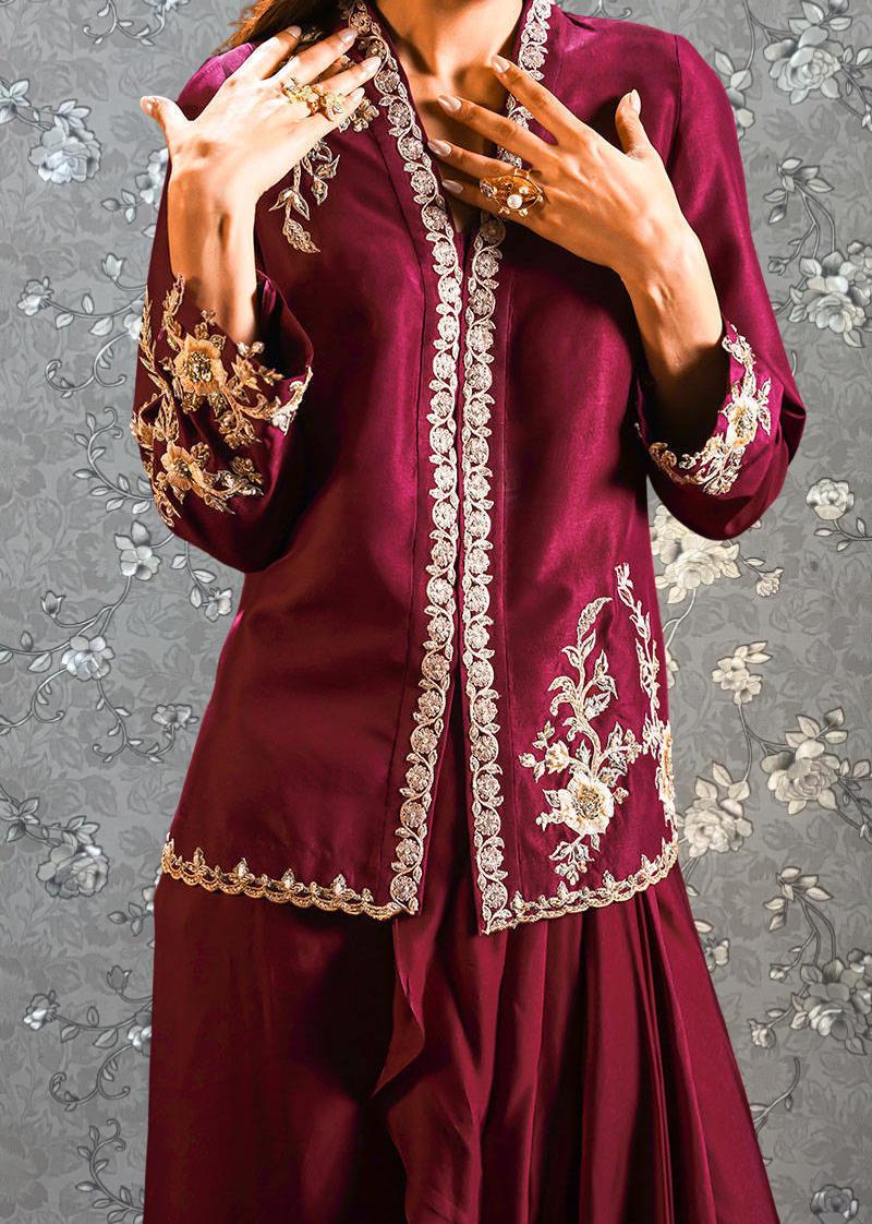 A woman wearing a burgundy dress with intricate embroidery and jewelry poses with her hands on her chest.