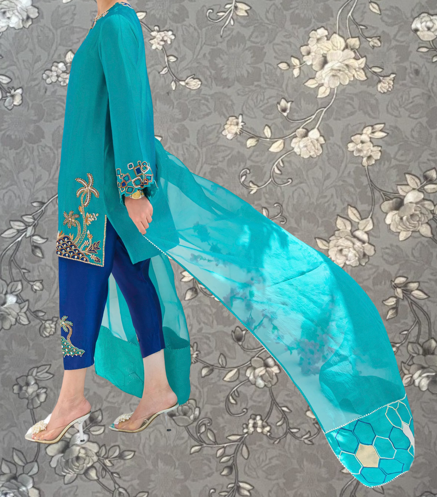 A person wearing a turquoise dress with intricate embroidery and a matching turquoise shawl is standing against a gray floral background.