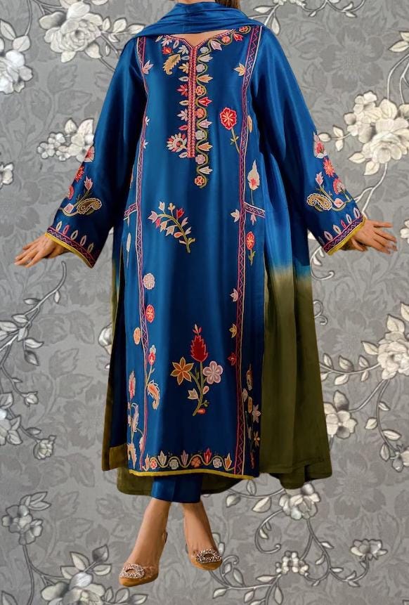 A woman wearing a long, blue dress with intricate floral embroidery stands against a gray floral background.