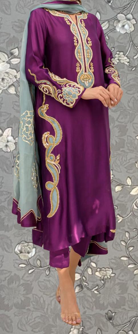 A person wearing a purple and gold dress with intricate embroidery and a matching scarf stands against a gray floral background.