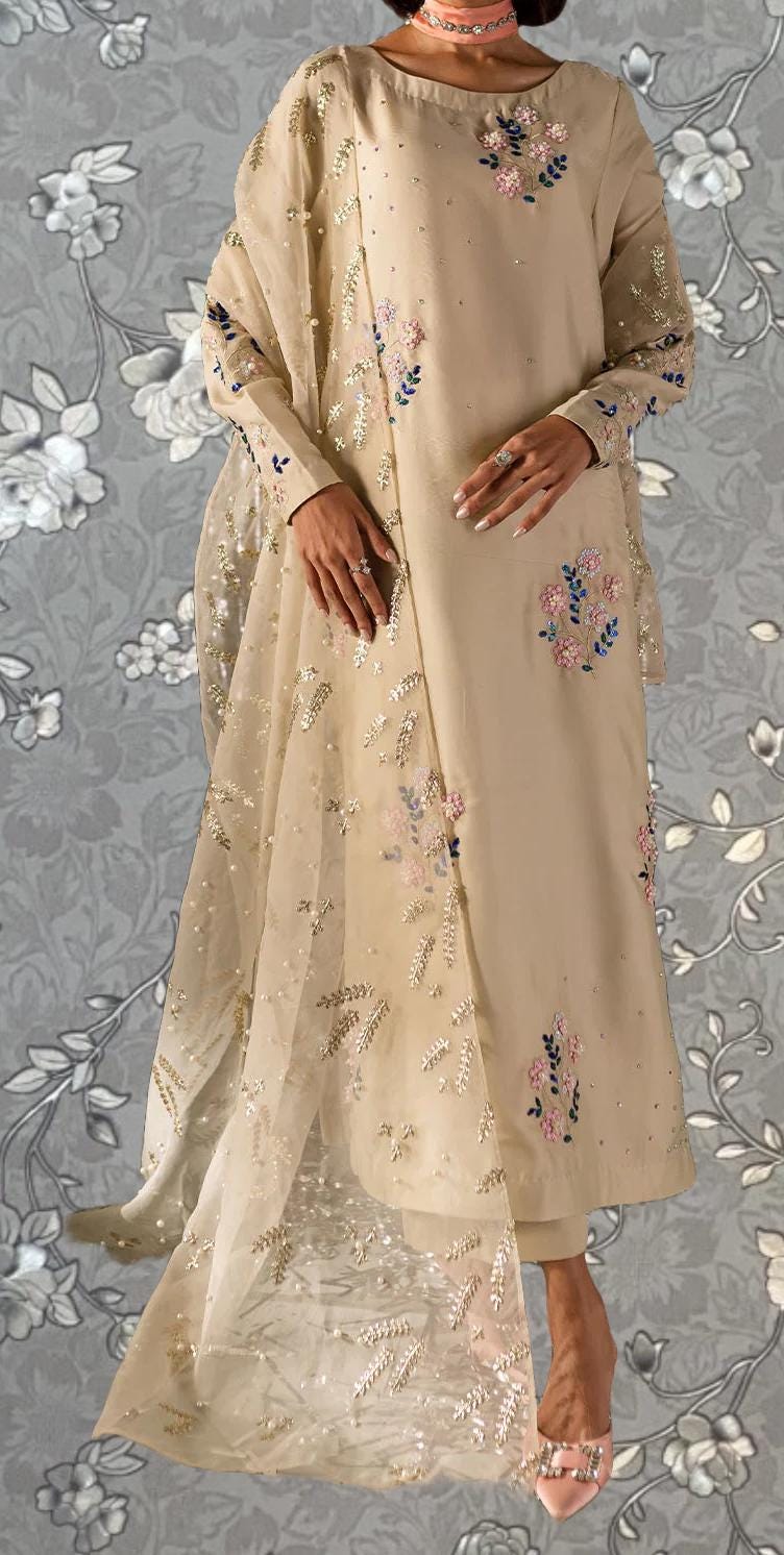 A woman wearing a beige dress with floral embroidery and a matching shawl stands against a gray floral background.