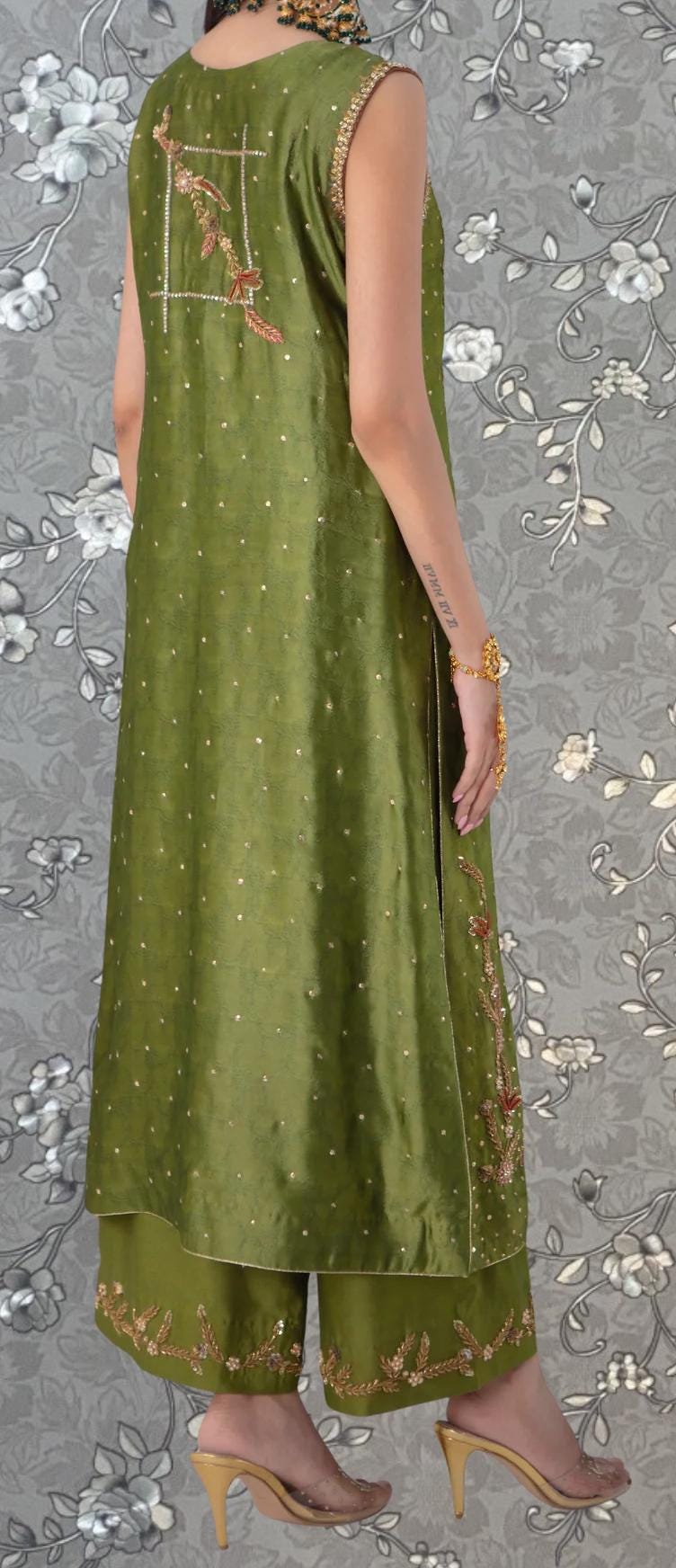 A person wearing a green dress with gold embroidery and gold heels is standing against a gray floral background.