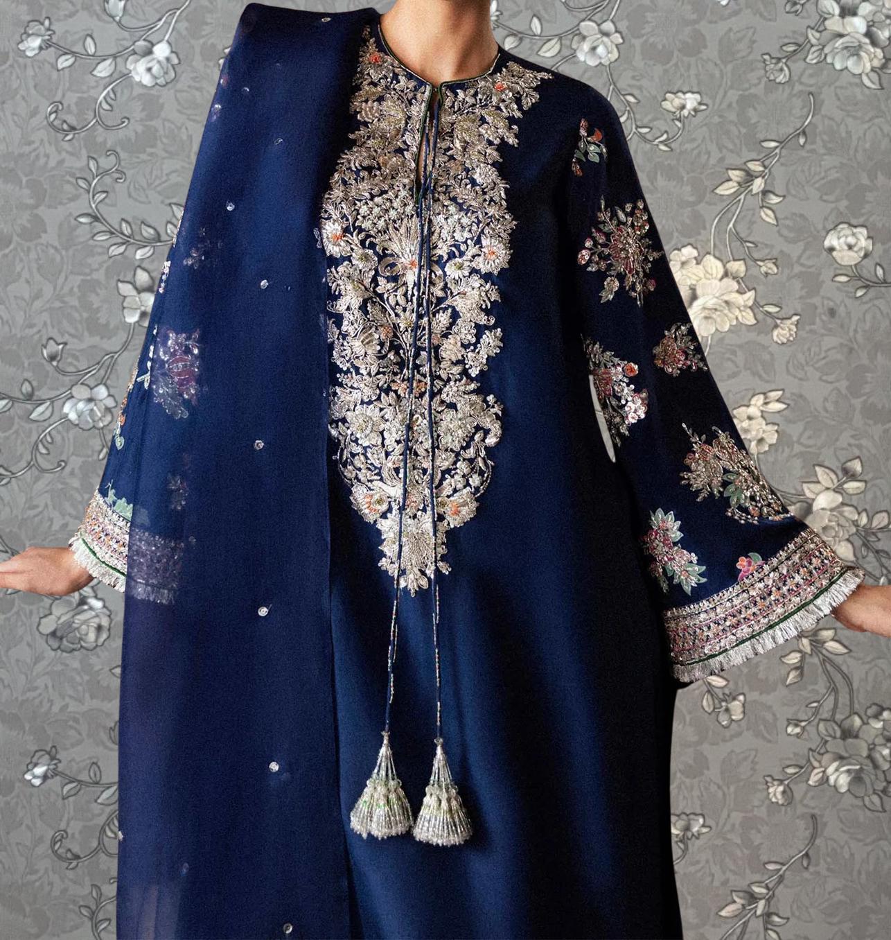 A woman wearing a blue dress with intricate embroidery and tassels, standing against a gray floral background.