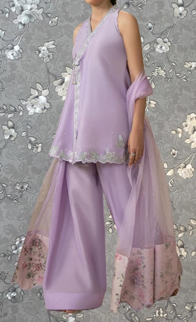 A woman wearing a lavender dress with a floral pattern and a matching shawl stands against a gray floral background.