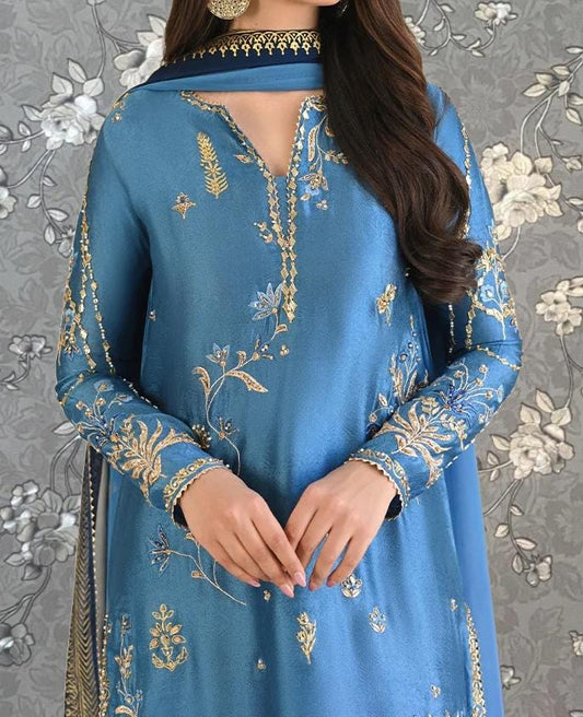 A woman wearing a blue dress with intricate gold embroidery poses against a gray floral background.