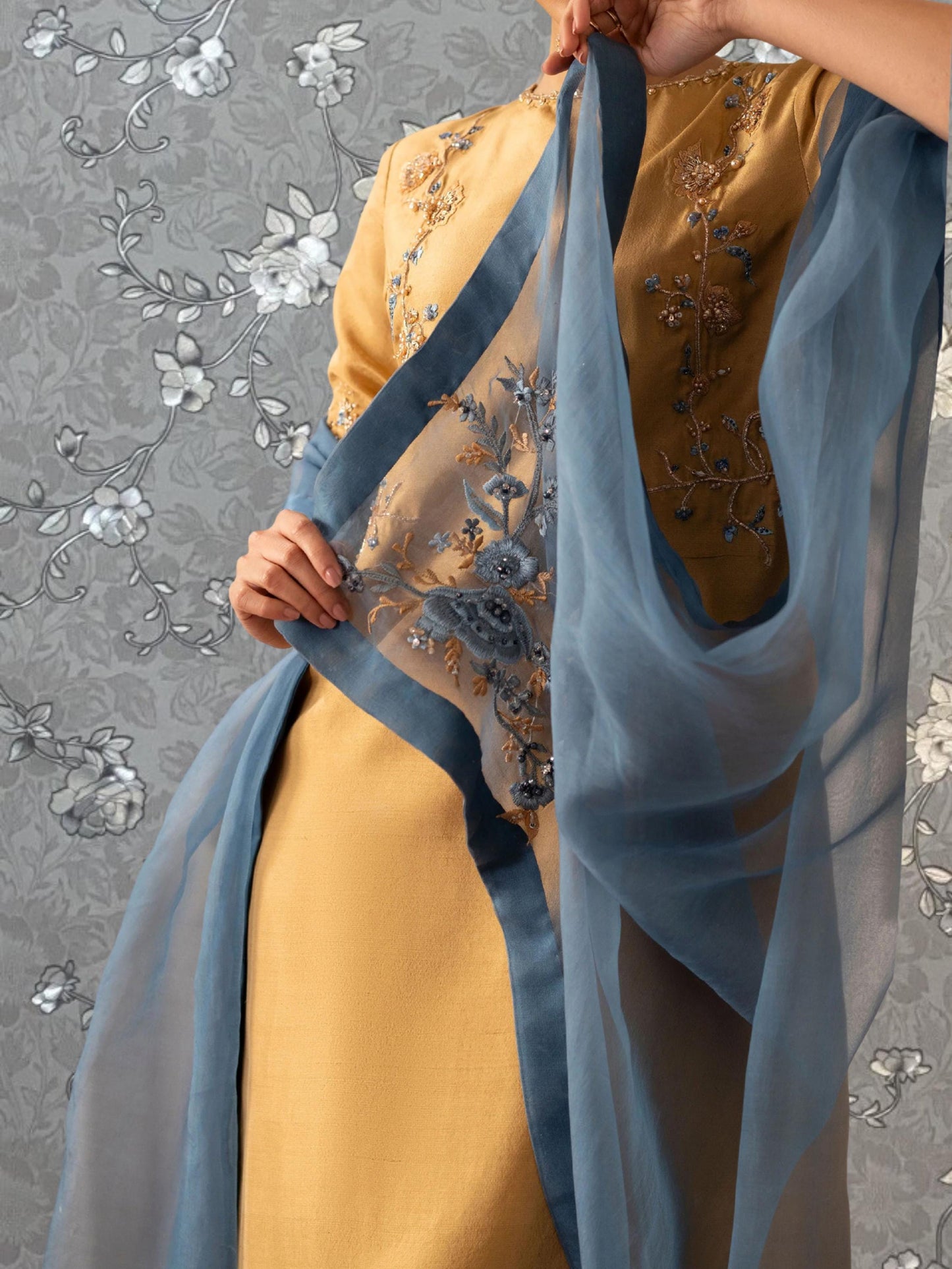 A person wearing a yellow and blue dress with intricate embroidery, holding a blue scarf.