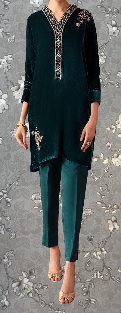 A person wearing a dark green velvet dress with gold embroidery and gold heels, standing against a gray floral background.