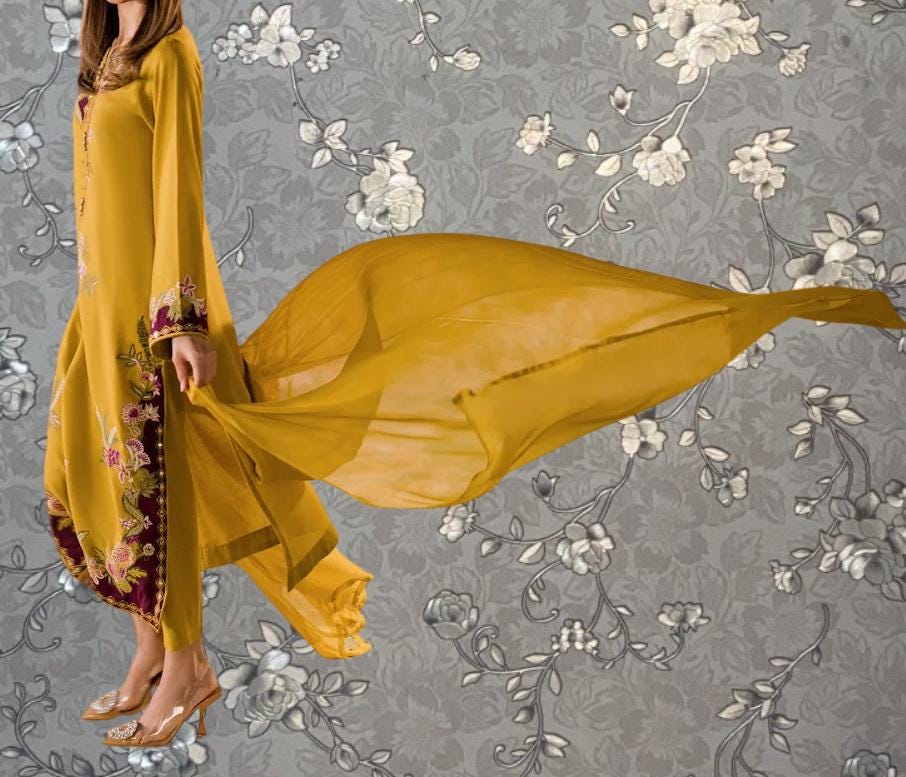 A woman wearing a long yellow dress with floral embroidery is standing against a gray floral background.