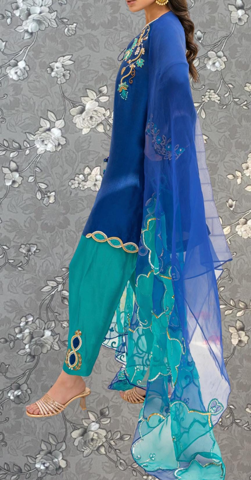 A woman wearing a blue and green dress with a long, flowing blue shawl, standing against a gray floral background.