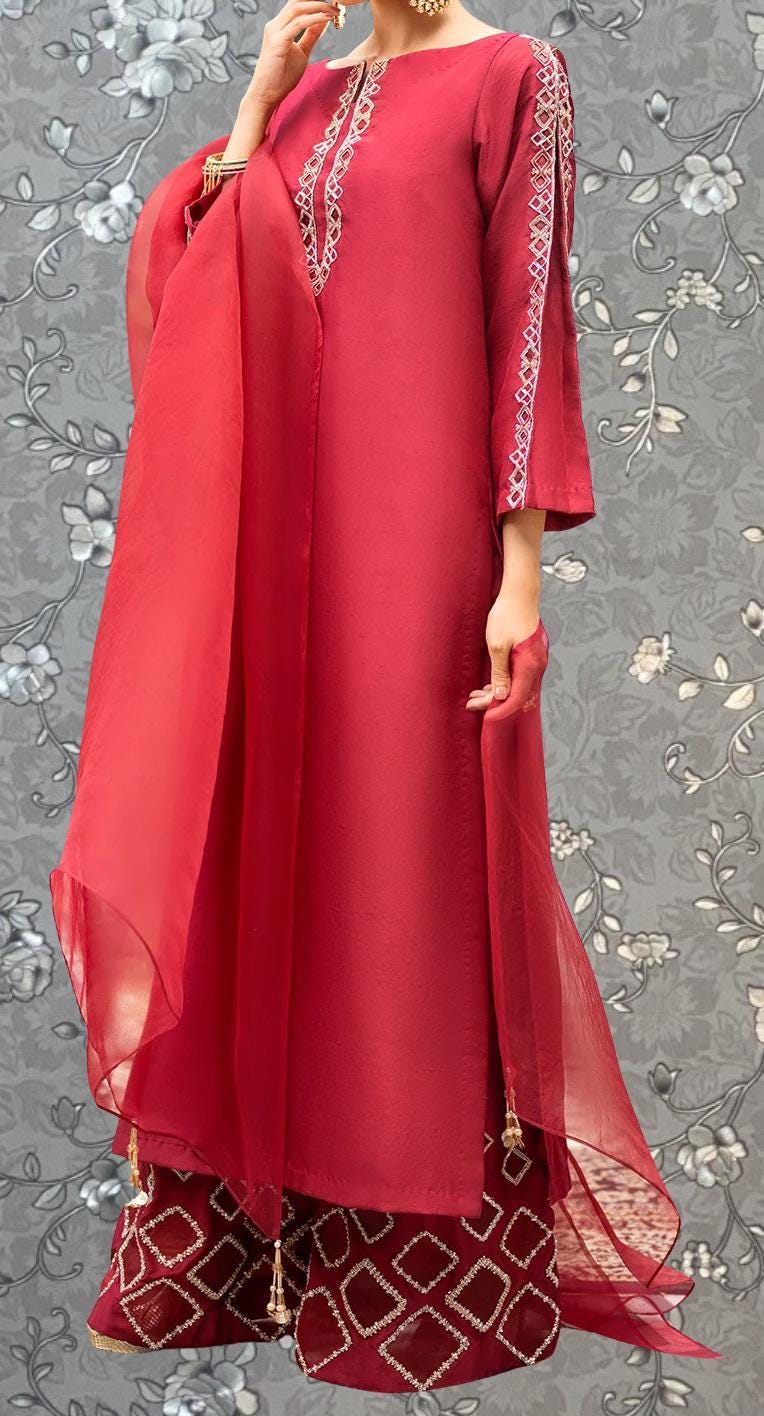 A woman wearing a long red dress with intricate embroidery and a matching red skirt, standing against a gray floral background.