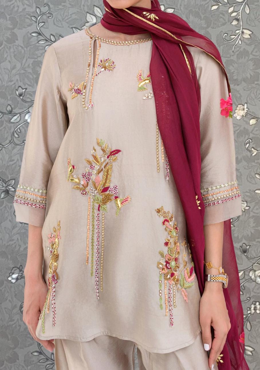 A person wearing a beige dress with intricate embroidered designs, including floral patterns and metallic accents. The dress is complemented by a burgundy scarf draped around the neck.