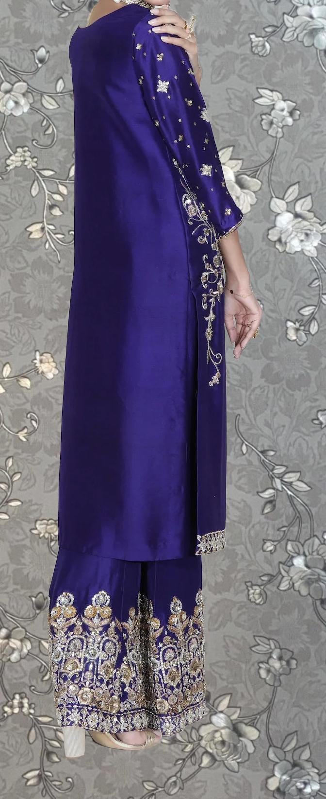 A person wearing a long, purple dress with gold embroidery on the sleeves and skirt, standing against a gray floral background.