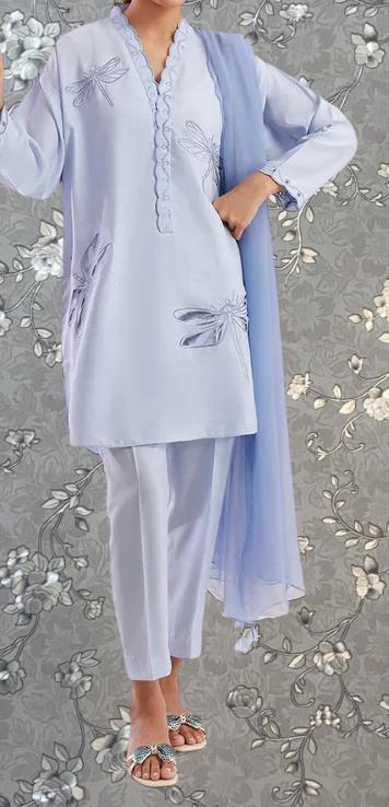 A person wearing a light blue dress with a pattern of blue dragonflies on it, standing against a gray floral background.