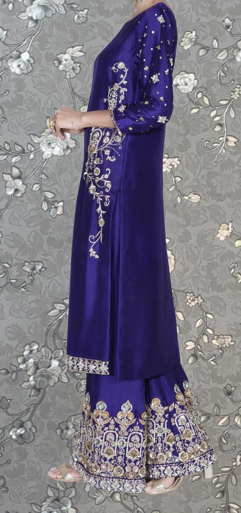 A woman wearing a long, purple dress with intricate gold embroidery stands against a gray floral background.