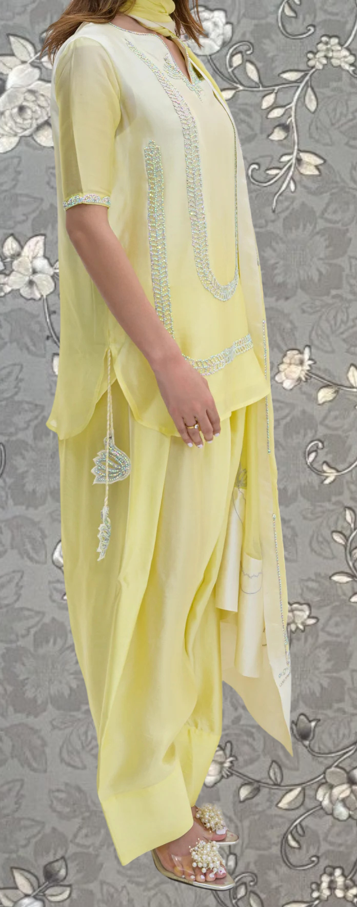 A woman wearing a yellow dress with a floral pattern and a matching yellow scarf is standing against a gray floral background.