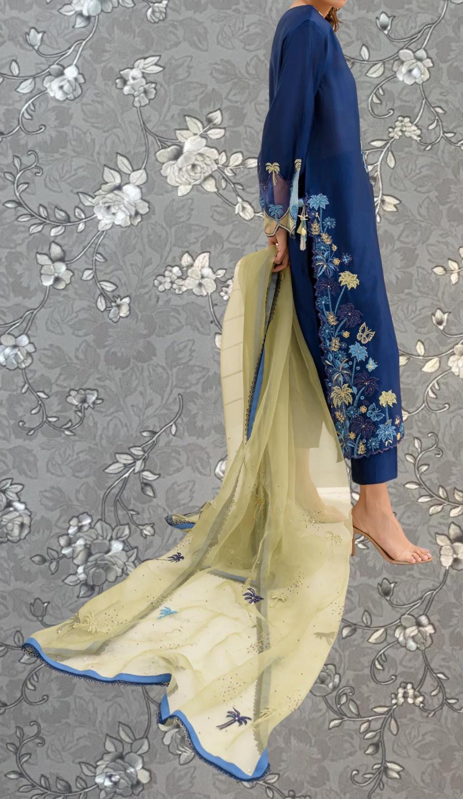 A person wearing a blue dress with intricate floral embroidery stands against a gray floral background.