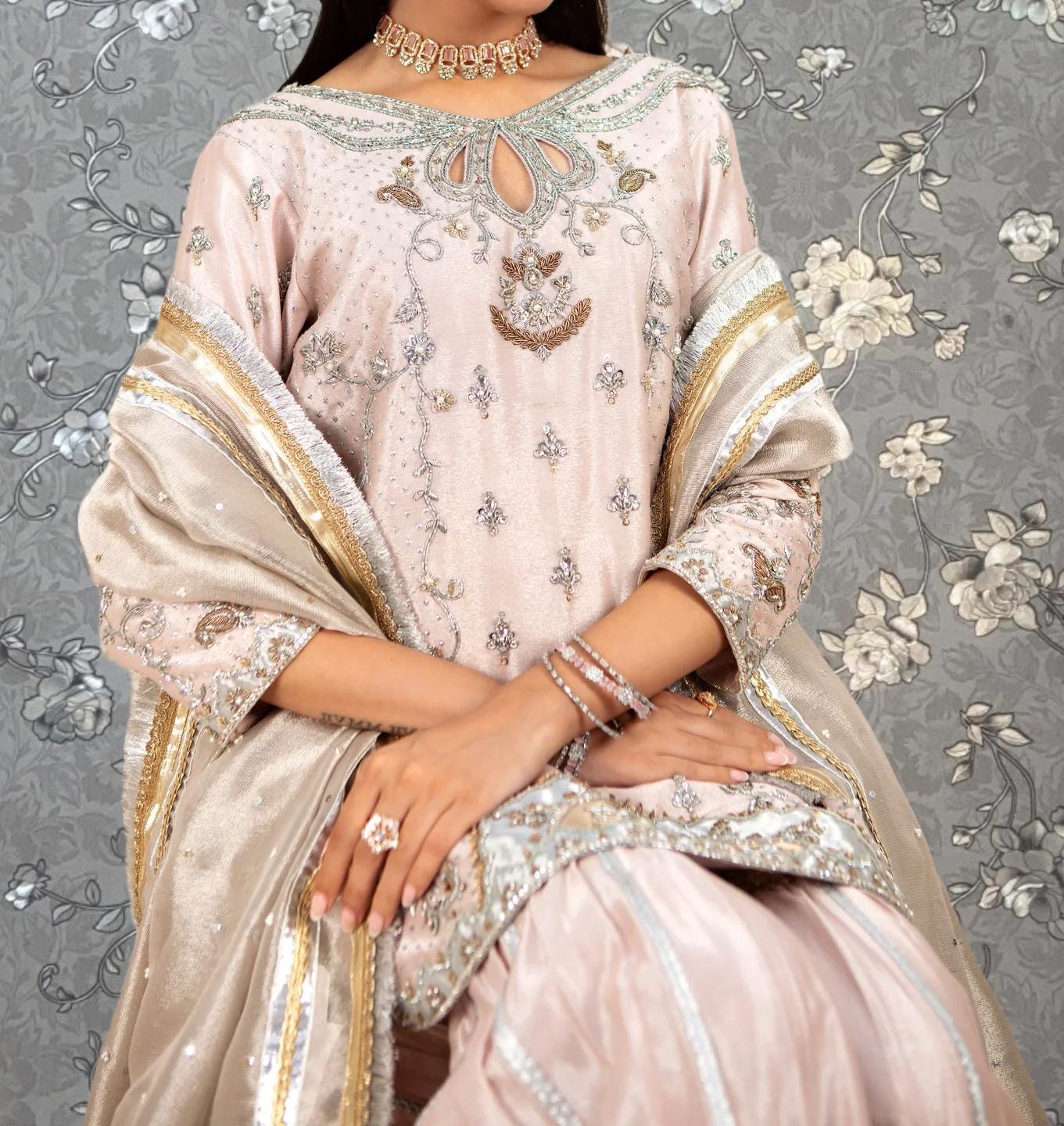 A woman wearing an ornate pink and gold dress with intricate embroidery and jewelry poses against a floral patterned background.