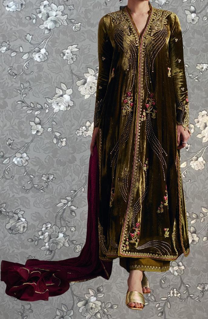 A person wearing a long, gold and burgundy velvet dress with intricate embroidery and floral patterns, standing against a gray floral background.