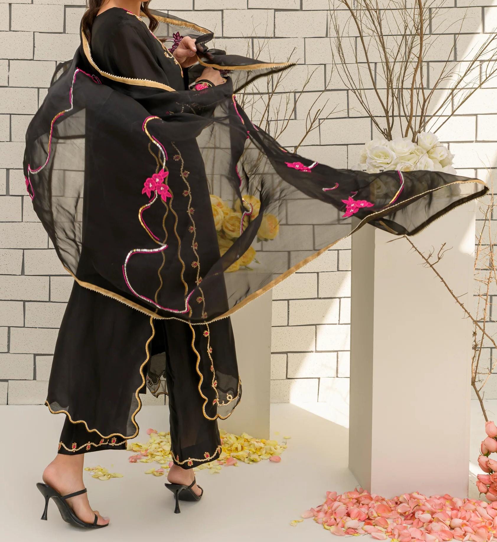 A woman in a black dress with pink accents stands in front of a white brick wall, holding a pink flower in her hand.