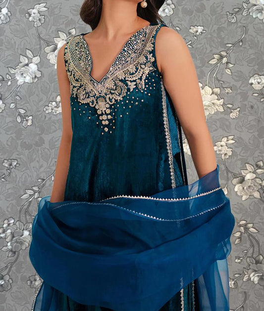 A woman wearing a blue dress with gold embroidery stands against a gray floral background.