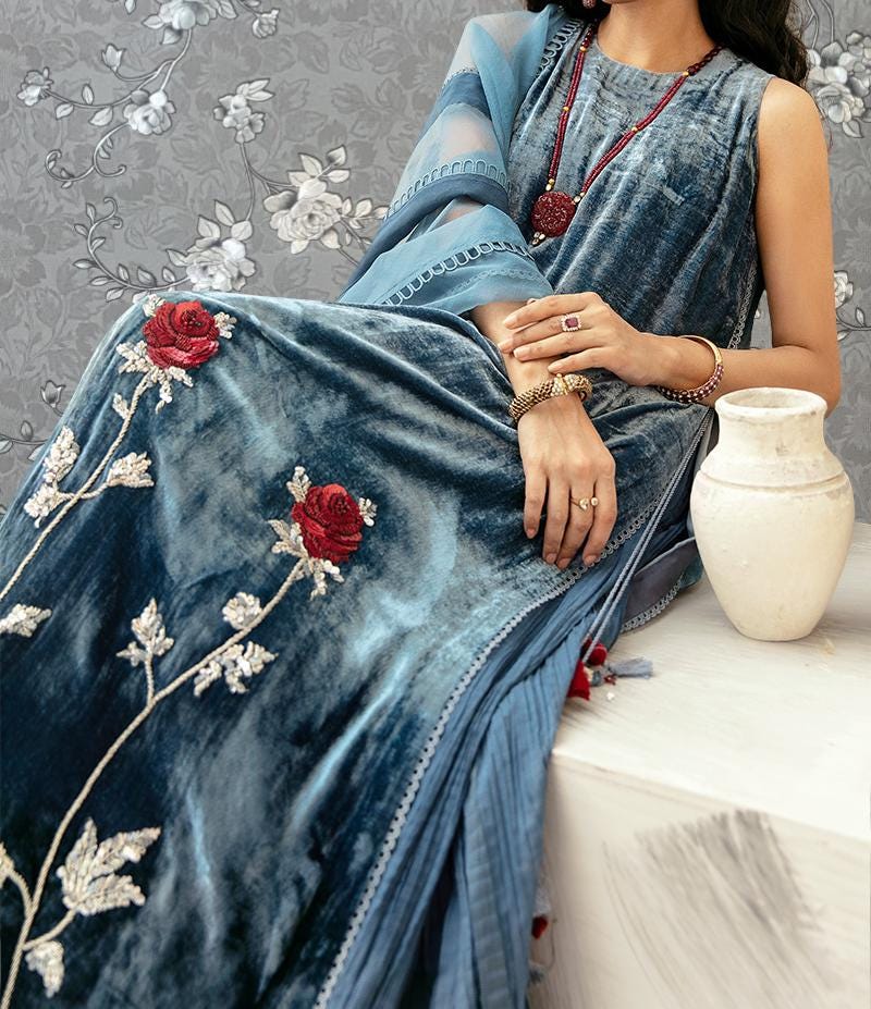 A woman wearing a blue velvet dress with red rose embroidery is sitting on a white surface next to a white vase.