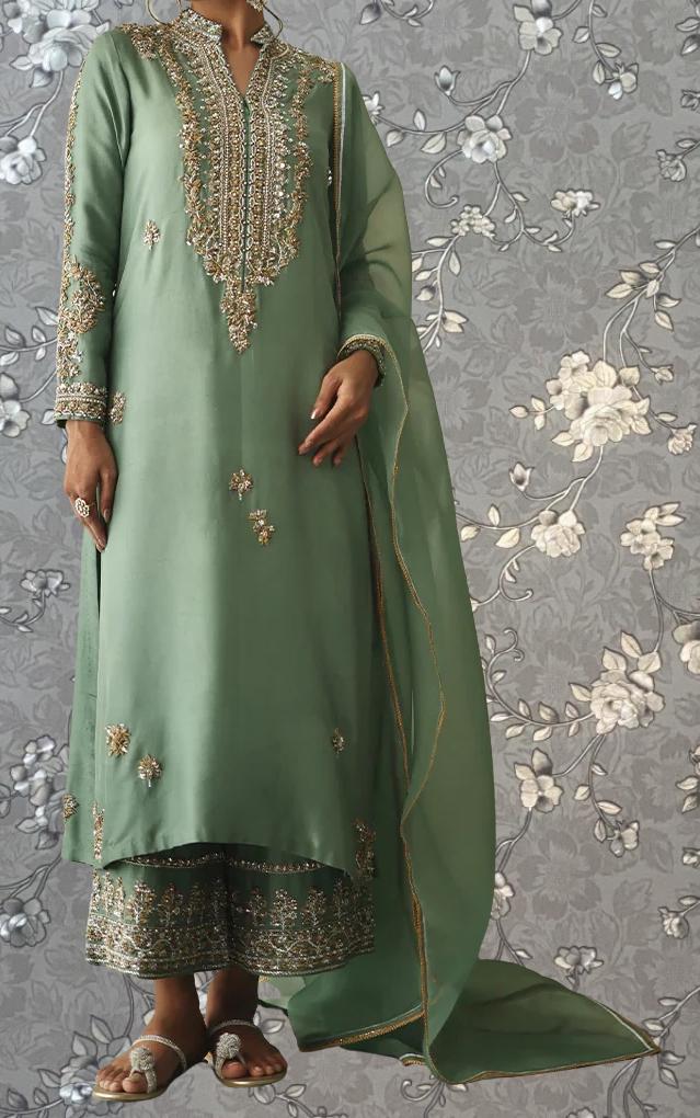 A person wearing a long, green dress with intricate embroidery and gold accents, standing against a gray floral background.