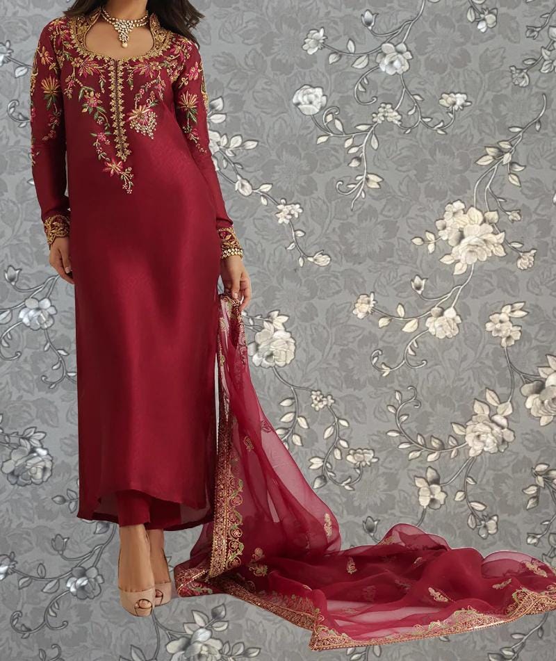 A woman wearing a long, red dress with intricate embroidery stands against a gray floral background.