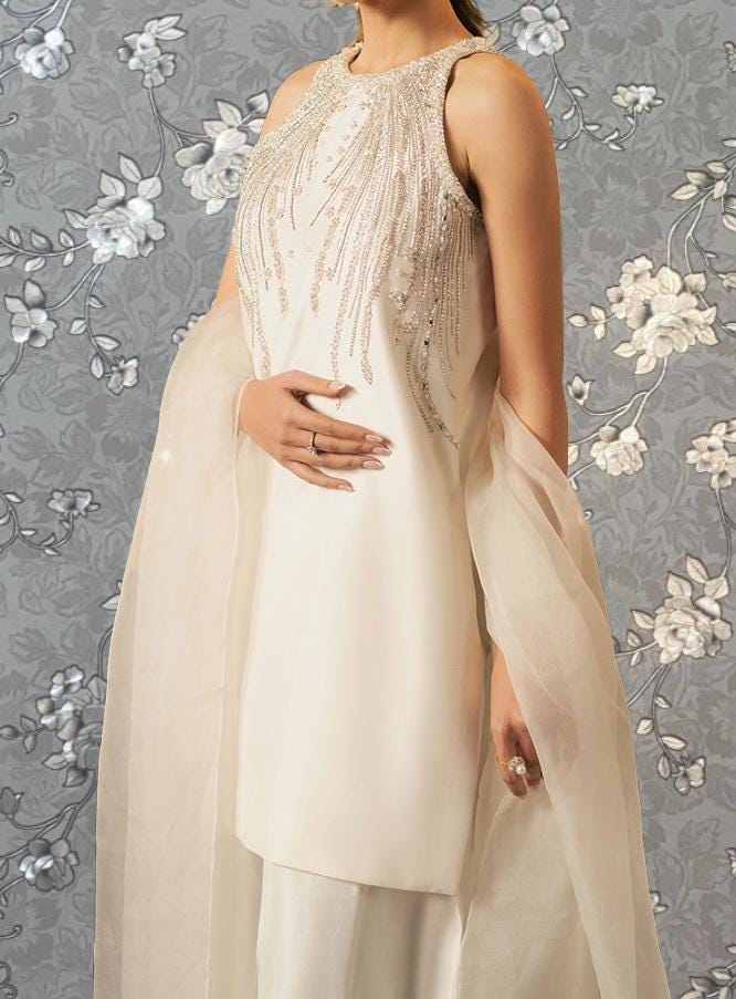 A woman wearing a white dress with a beaded bodice and sheer fabric draped over her shoulders, standing against a gray floral background.