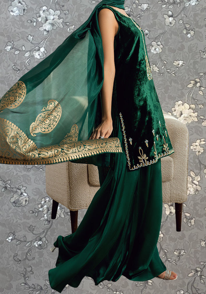 A woman wearing a green velvet dress with gold embroidery and a matching green shawl is standing in front of a gray floral background.