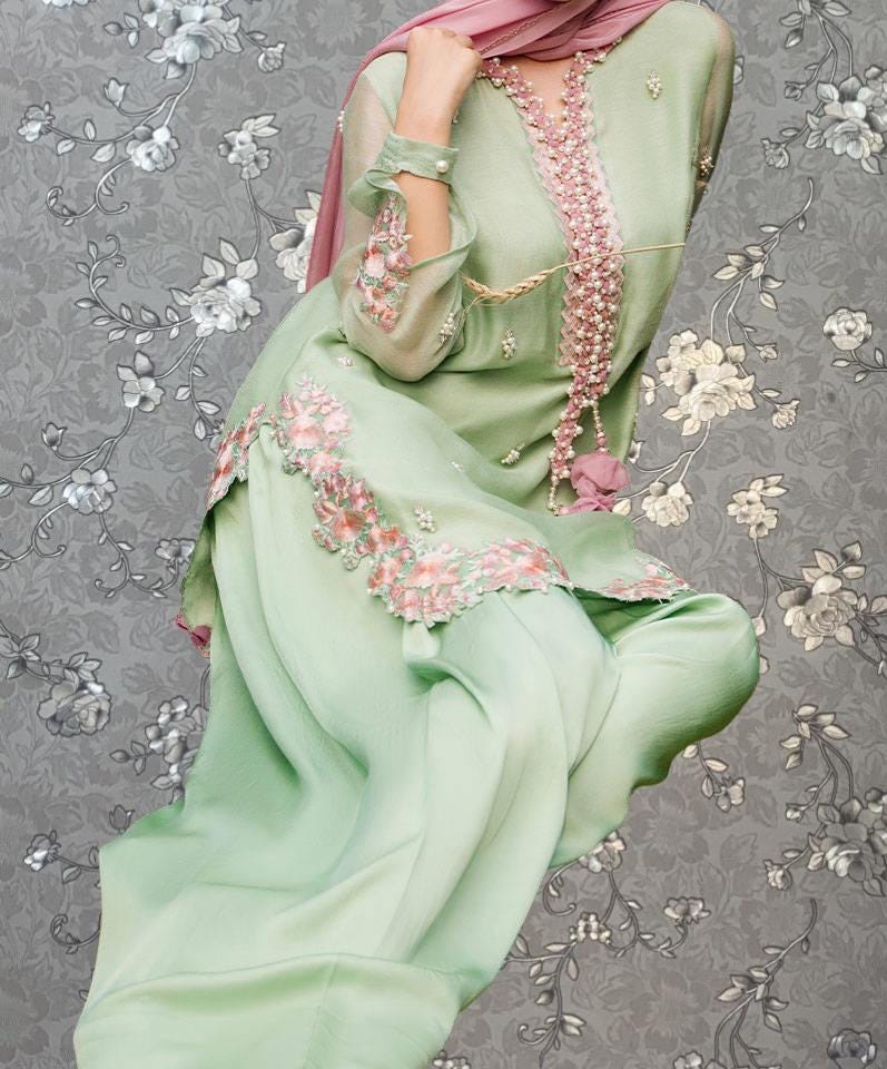 A woman wearing a green dress with pink floral embroidery and a pink scarf is shown against a gray floral background.