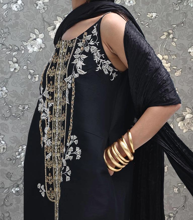A woman wearing a black dress with gold embroidery and gold bracelets stands against a gray floral background.