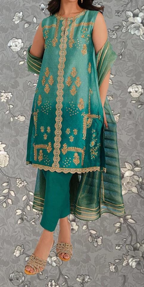 A woman wearing a green and gold dress with intricate embroidery and patterns, standing against a gray floral background.