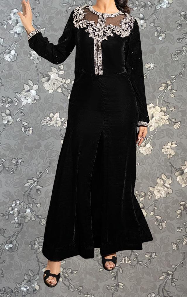 A woman wearing a long black dress with a floral pattern on the bodice and skirt, posing against a gray floral background.