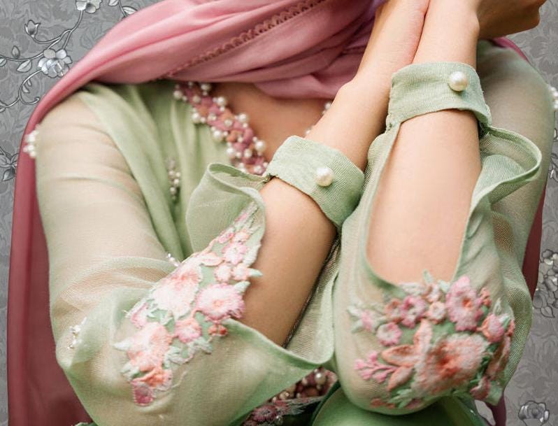 A close-up of a person's hands wearing a green dress with pink floral embroidery, and a pink scarf draped over their shoulders.