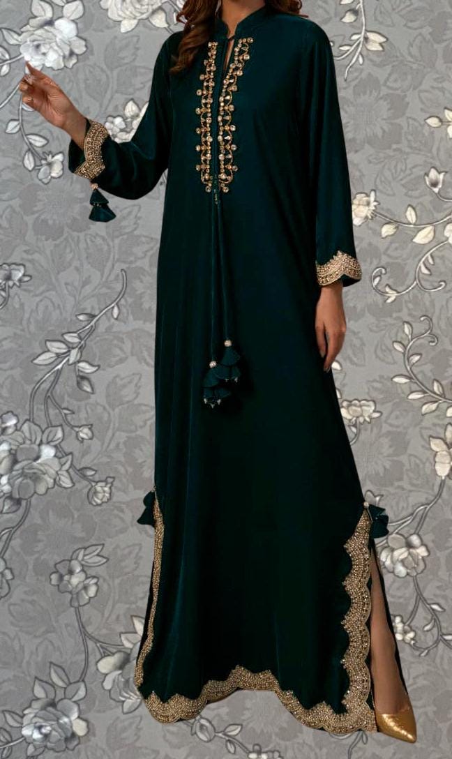 A woman wearing a long, dark green dress with gold embroidery and tassels, standing against a gray floral background.